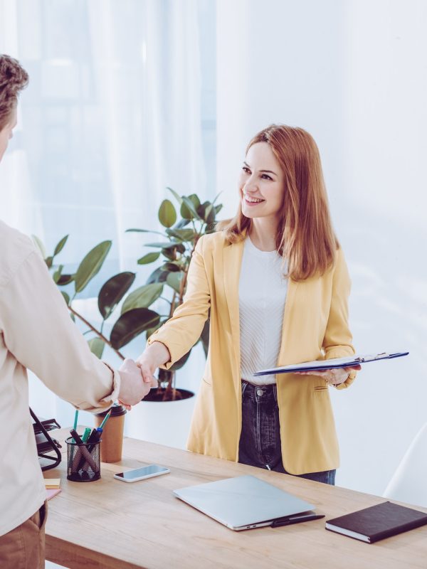 happy-recruiter-holding-clipboard-and-shaking-hands-with-employee-in-modern-office.jpg